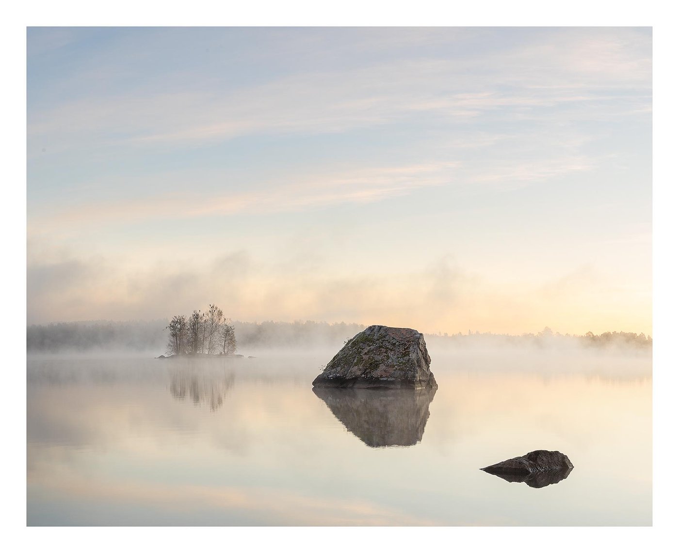 A misty lake at sunrise with rocks and a small wooded island reflected in still water